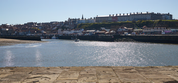 This landscape photograph captures the Yorkshire coastal town of Whitby on a sunny spring afternoon. The image is taken from the bay, with the water shimmering in the bright sunlight, and numerous boats can be seen in the harbor. The main subject of the photo is the picturesque town of Whitby, with its distinctive architecture, including rows of houses and prominent historic buildings on the hill. Whitby’s iconic skyline features landmarks such as the grand Royal Hotel and the spire of St. Mary’s Church rising above the town. The scene conveys the charm and character of this Yorkshire destination, with its mix of seaside and urban elements, nestled against the backdrop of the bay and cliffs.